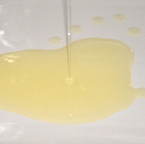 Hellgelbes, kaltgepresstes Mandelöl, das aus einem Glas auf helles Leinen fließt – natürliches Basis- und Trägeröl für empfindliche Haut